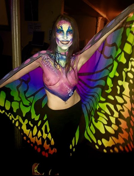 A photo of a woman with her torso painted to look like she is wearing a transparent shirt, and wearing a colorful cape that looks like butterfly wings as she spreads her arms.